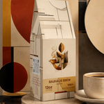 A 12 oz package of Bauhaus Brew coffee stands on a round pedestal, styled with scattered coffee beans and a cup of coffee on a saucer against an abstract geometric background.
