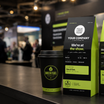 Custom coffee packaging mockup on a dark countertop, featuring a black and neon yellow-green coffee cup, a matching coffee bag, and a coffee box with placeholder branding and event text, set against a blurred convention background.