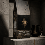A bag of Brutalist Blend coffee sits on a concrete pedestal in a dark, minimalist industrial setting, with a black ceramic pot beside it.