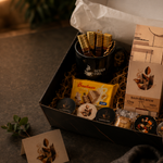 An open gift box filled with assorted packaged snacks and coffee items, photographed on a tabletop with a person's hands opening the lid. Visible products include a black cup with wrapped sticks, a yellow packet, round chocolate or candy pieces, and a tall beige box labeled 'MADAGASCAR BEWAF'.