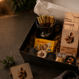 An open gift box filled with assorted packaged snacks and coffee items, photographed on a tabletop with a person's hands opening the lid. Visible products include a black cup with wrapped sticks, a yellow packet, round chocolate or candy pieces, and a tall beige box labeled 'MADAGASCAR BEWAF'.