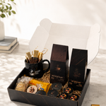 A gourmet gift box set displayed on a marble surface with a white background. The set includes a black coffee mug with gold-foiled straws, two tall black boxes of coffee, packaged snacks, and a few loose chocolates.