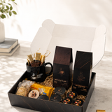 A gourmet gift box set displayed on a marble surface with a white background. The set includes a black coffee mug with gold-foiled straws, two tall black boxes of coffee, packaged snacks, and a few loose chocolates.
