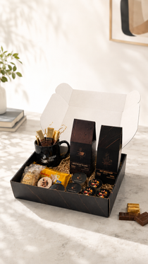 A gourmet gift box set displayed on a marble surface with a white background. The set includes a black coffee mug with gold-foiled straws, two tall black boxes of coffee, packaged snacks, and a few loose chocolates.