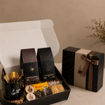 A coffee gift set displayed in an open black presentation box on a beige surface, alongside a closed matching gift box with a ribbon. The set includes packaged coffee bags, a small gold-toned cup, assorted snacks, and a branded card, arranged against a neutral beige background.