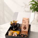 A gourmet gift box filled with assorted chocolates, a black coffee mug with gold-wrapped sticks, and packaged snacks, displayed on a light marble surface with a green plant in the background.