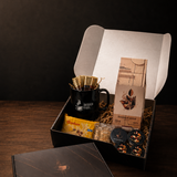 A curated gift box set displayed on a dark wooden table against a black background. The set includes a black coffee mug with a gold filter, a box of coffee beans, a small yellow packet, and a round tin, alongside a black presentation box lid in the foreground.
