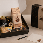 A coffee-themed gift box set displayed on a tabletop, featuring an open black box with packaged coffee accessories, a closed matching black gift box with a brown ribbon, a black mug, a bowl of coffee beans, and a pen on a neutral beige background.