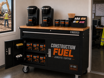 A black rolling utility coffee cart with a wood-look top, branded Construction Fuel, shown in an office setting with its door open to reveal stocked coffee bags. Two black coffee makers and matching mugs sit on the counter.