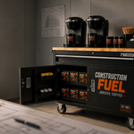 A black rolling coffee cart with a wood top in a construction office setting. The cart has 'CONSTRUCTION FUEL JOBSITE COFFEE' printed on the side, with an open door showing packaged coffee supplies inside.