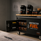 A black rolling coffee cart with a wood top in a construction office setting. The cart has 'CONSTRUCTION FUEL JOBSITE COFFEE' printed on the side, with an open door showing packaged coffee supplies inside.