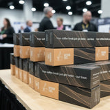 Stacked black and tan boxes of Kevrin Pro coffee break boxes on a wooden table at a busy trade show or convention, with blurred people and booths in the background.