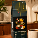 A bag of Mid-Mod Morning Light 100% roasted coffee sits on a wooden table in a warm, modern room setting, next to a ceramic cup of coffee and scattered coffee beans.