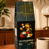 A bag of Mid-Mod Morning Light 100% roasted coffee sits on a wooden table in a warm, modern room setting, next to a ceramic cup of coffee and scattered coffee beans.