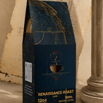 A dark navy coffee bag labeled 'RENAISSANCE ROAST' stands on a tabletop with vintage props, including an aged paper sketchbook, a column base, and a brass compass. The package design features gold celestial line art and text indicating 100% roasted coffee and 12oz size.