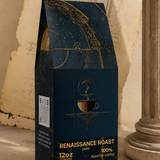A dark navy coffee bag labeled 'RENAISSANCE ROAST' stands on a tabletop with vintage props, including an aged paper sketchbook, a column base, and a brass compass. The package design features gold celestial line art and text indicating 100% roasted coffee and 12oz size.